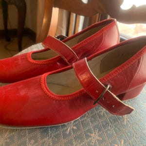 May include: A pair of red leather Mary Jane style shoes with a strap and buckle closure. The shoes have a rounded toe and a small heel. The shoes are sitting on a patterned surface.