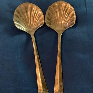 May include: Two antique gold-colored serving spoons. The spoon bowls are shaped like scallop shells with detailed ridged patterns. The handles are long and slender. The spoons are displayed against a dark blue background.