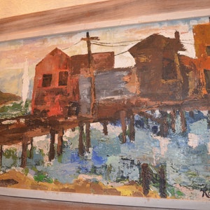 May include: An abstract oil painting of a waterfront scene with buildings in shades of red, brown, and orange. The buildings are reflected in the water below. The painting is framed in a light-colored wooden frame.