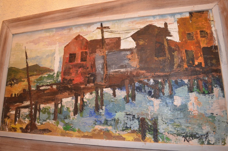 May include: An abstract oil painting of a waterfront scene with buildings in shades of red, brown, and orange. The buildings are reflected in the water below. The painting is framed in a light-colored wooden frame.