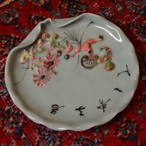 May include: A light blue ceramic dish with a raised floral design in shades of pink, green, and white. The dish has a scalloped edge and features black calligraphy writing on the bottom.