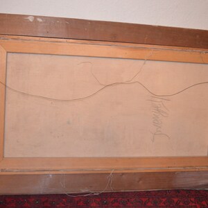 May include: The back of a framed painting with a light brown wooden frame. The canvas is a light beige color. The frame has a wire for hanging. The artist's signature is visible in pencil on the canvas.