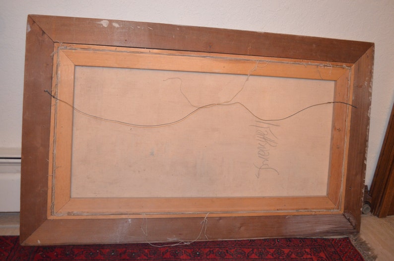 May include: The back of a framed painting with a light brown wooden frame. The canvas is a light beige color. The frame has a wire for hanging. The artist's signature is visible in pencil on the canvas.