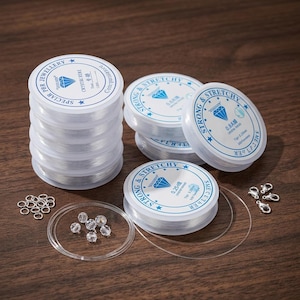 May include: A collection of clear elastic jewelry cord spools with white labels featuring blue text and diamond icons. The set includes several spools of varying thicknesses, small silver jump rings, silver lobster clasps, and clear beads.