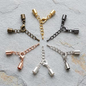 Könnte beinhalten: Eine Sammlung von sechs mehrsträngigen Schmuckverbindern in verschiedenen metallischen Ausführungen wie Gold, Silber, Roségold, Gunmetal und Bronze. Jeder Verbinder verfügt über zwei zylindrische Endkappen und einen Karabinerverschluss.
