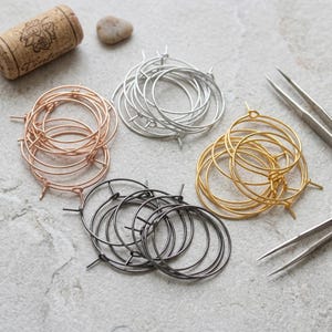 May include: A collection of circular metal hoop earrings in four distinct finishes: rose gold, silver, gold, and gunmetal grey. Each set consists of multiple thin wire hoops with a simple clasp mechanism, arranged in clusters on a textured surface.