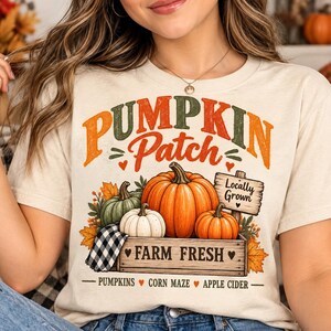 May include: A cream-colored t-shirt featuring a graphic with the text PUMPKIN PATCH, LOCALLY GROWN, FARM FRESH, PUMPKINS, CORN MAZE, and APPLE CIDER. The design includes orange, white, and green pumpkins, autumn leaves, and a wooden crate.