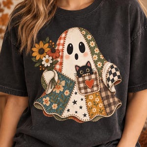 May include: A charcoal gray t-shirt featuring a large graphic of a patchwork ghost holding a bouquet of sunflowers and daisies. The ghost has a small black cat peeking out of a pocket with a red heart, surrounded by various floral and star patterns.