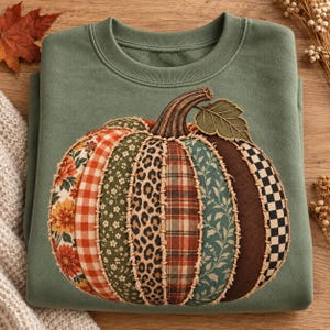 May include: A sage green crewneck sweatshirt featuring a large, central pumpkin graphic. The pumpkin is composed of vertical fabric patches with diverse patterns, including floral, orange gingham, leopard print, plaid, and black and white checks.