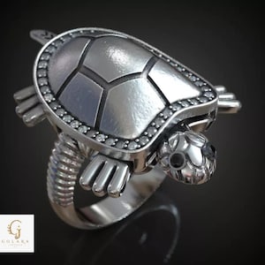 May include: A silver-toned ring featuring a turtle design with a smooth, segmented shell, small clear stones bordering the shell, and dark stones set as eyes. The band has a ribbed texture, and the logo GOLARA JEWELRY is visible in the corner.