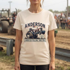 May include: A cream-colored short-sleeve t-shirt featuring a graphic of a modified pulling tractor. The text reads ANDERSON above the tractor and 74 MODIFIED PULLING TRACTOR below it. The design includes a blue and red tractor with large rear tires.