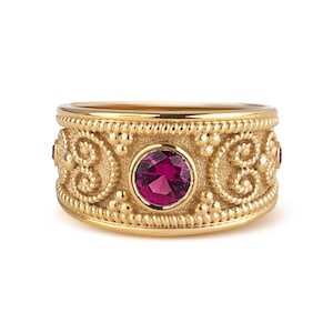 May include: A wide gold band ring featuring a central round-cut magenta gemstone set in a bezel. The surface of the ring is decorated with intricate granulated metalwork, including beaded borders and swirling scroll patterns in a textured finish.