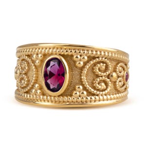May include: A wide gold band ring featuring a central oval-cut deep pink gemstone. The surface of the ring has a textured, matte finish with raised beaded edges and intricate scrollwork patterns surrounding the central stone setting.