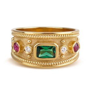 May include: A gold band ring featuring a central emerald-cut green gemstone, flanked by two round white diamonds and two round red rubies. The ring has a textured matte finish with rope-style edges and small decorative gold spheres.