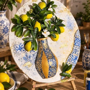 May include: A circular mixed-media art piece on a wooden easel featuring a three-dimensional vase with blue and white patterns and gold accents. The vase holds a branch with yellow lemons, green leaves, and white flowers against a textured white background.