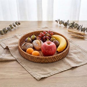 May include: A round, woven rattan tray filled with a variety of fresh produce, including a bright red apple, a yellow banana, a cluster of purple grapes, two small orange apricots, two dark purple figs, and two small, round, textured brown loaves.