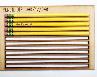 Puede incluir: Un jig de madera para lápices con 12 ranuras para lápices. El jig está etiquetado como "PENCIL JIG 340/12/340". Hay cuatro lápices amarillos en las ranuras, con un lápiz etiquetado como "Go Ravens!"