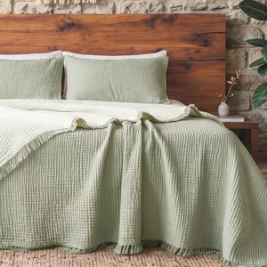 May include: A sage green bedspread with a textured waffle weave pattern and fringed edges, paired with two matching rectangular pillows. The bedding is draped over a bed frame featuring a dark wood headboard against a stone wall background.