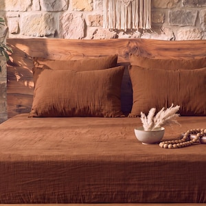 May include: A bed set featuring a solid rust-colored cotton muslin flat sheet and two matching rectangular pillowcases. A small ceramic bowl containing dried pampas grass and a strand of wooden beads are placed on the bed surface.