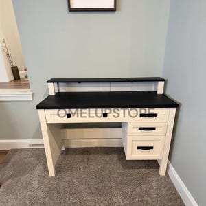May include: A white desk with a black top and a matching black hutch shelf. The desk features three drawers on the right side and one wide drawer in the center, all equipped with black handles. The text OMELUPSTORE is visible on the front.
