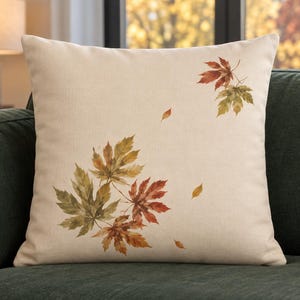 May include: A square throw pillow with a light beige, textured fabric cover. The front features a printed design of scattered maple leaves in shades of green, yellow, orange, and deep red, arranged in two clusters with smaller individual leaves.