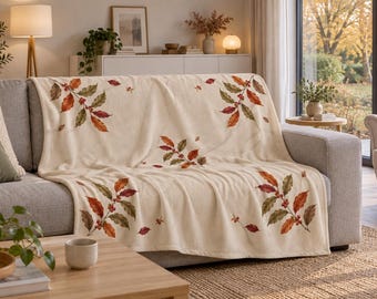 May include: A cream-colored throw blanket draped over a sofa, featuring a repeating pattern of autumn-toned leaves in shades of orange, red, and brown, interspersed with small clusters of red berries across the fabric surface.