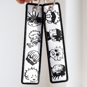May include: Two rectangular fabric keychains featuring black and white embroidered anime-style character faces. Each keychain has a silver metal ring attached through a reinforced grommet at the top, with black stitched borders on a white background.
