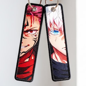 Peut inclure: Deux porte-clés rectangulaires en tissu avec des illustrations de personnages d'anime. Le porte-clés de gauche montre un personnage aux cheveux rouges, yeux rouges et marques sombres. Celui de droite a des cheveux blancs et yeux bleus.