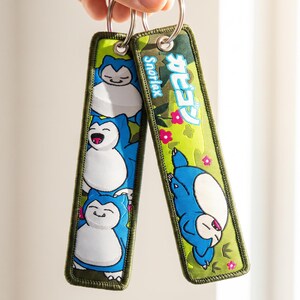 Puede incluir: Dos llaveros rectangulares de tela con bordados de personajes Snorlax azules y crema sobre un fondo de camuflaje verde. Un llavero muestra el texto Snorlax y caracteres japoneses en blanco y azul, acentuado con pequeñas flores rosas.