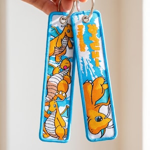 Könnte beinhalten: Zwei rechteckige Schlüsselanhänger aus Stoff mit blauen Rändern und Metallringen. Einer zeigt drei orangefarbene drachenartige Kreaturen, der andere eine orangefarbene Kreatur sowie den vertikalen Text "Dragonite" und japanische Schriftzeichen.