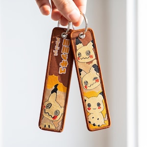 Può includere: Due portachiavi rettangolari in tessuto marrone con personaggi Mimikyu ricamati. Un portachiavi mostra il testo giapponese "ミミッキュ" e quello inglese "Mimikyu" in verticale, mentre l'altro presenta tre volti di Mimikyu sovrapposti con dettagli neri e rossi.