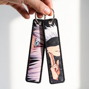 Op de afbeelding: Twee rechthoekige stoffen sleutelhangers met anime-stijl karakterillustraties. De ene toont een close-up van een blauw oog en wit haar, de andere een personage met een zwarte blinddoek en wit haar dat een handgebaar maakt.