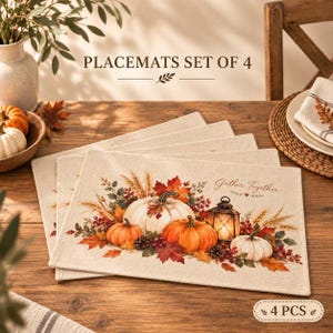 May include: A set of four rectangular placemats featuring a printed autumn design of white and orange pumpkins, red maple leaves, wheat stalks, and a lit lantern. The text "Gather Together" is printed in script, and "PLACEMATS SET OF 4" is visible.