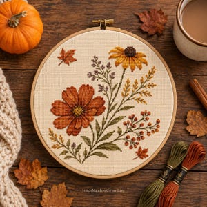 Op de afbeelding: Een ronde houten borduurring met een kruissteekpatroon van herfstbloemen, waaronder een grote oranje kosmos en een gele zonnehoed, met kleine blaadjes en bessen op crèmekleurige stof. De tekst StitchMeadowCo on Etsy is zichtbaar.