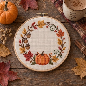 May include: A circular wooden embroidery hoop containing a cross-stitch pattern of an autumn wreath. The design features a central orange pumpkin surrounded by colorful red, yellow, and orange maple leaves, brown acorns, and green swirling vines.