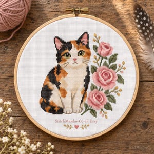 Peut inclure: Un tambour à broder circulaire en bois contenant un motif au point de croix représentant un chat calico assis à côté de trois roses roses avec des feuilles vertes. Le texte StitchMeadowCo on Etsy est imprimé sous le chat avec un petit cœur.