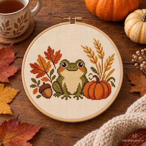May include: A circular wooden embroidery hoop containing a cross-stitch design of a green frog centered between autumn leaves, an acorn, a pumpkin, and stalks of wheat on a cream-colored fabric background.