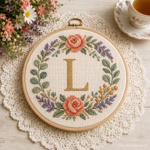 Pode incluir: Um bastidor de bordado circular de madeira contendo um desenho em ponto cruz sobre tecido branco sujo. O design apresenta uma letra L dourada grande no centro, rodeada por uma coroa de flores de rosas cor-de-rosa, lavanda roxa e folhas verdes.