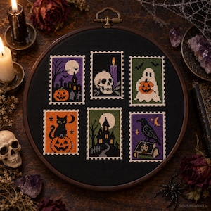 May include: A circular wooden embroidery hoop containing black fabric with six cross-stitched rectangular designs featuring Halloween motifs: a haunted house, a skull with a candle, a ghost with a pumpkin, a cat on a pumpkin, a house, and a raven on a book.