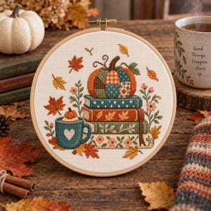 Könnte beinhalten: Ein runder Stickrahmen mit einem Kreuzstichmuster aus einem Patchwork-Kürbis auf einem Stapel von drei Büchern, neben einer blaugrünen Tasse mit Herzmotiv und einem kleinen Kürbis, umgeben von herbstlichen Blättern und floralen Zweigen.