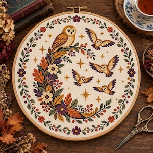 Op de afbeelding: Een ronde borduurring met een kruissteekpatroon van een grote uil gezeten op een halvemaanvormige krans van herfstbladeren, bessen en bloemen, omringd door vier kleinere vliegende uilen en gouden sterren op een crèmekleurige ondergrond.