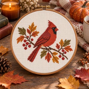 May include: A circular wooden embroidery hoop containing a cross-stitch pattern of a red cardinal bird perched on a branch with autumn leaves and red berries. The design is stitched on white fabric, featuring shades of orange, yellow, and green.