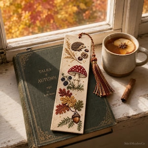 May include: A rectangular fabric bookmark featuring cross-stitch embroidery of a hedgehog, a red and white mushroom, autumn leaves, and an acorn. The bookmark is finished with a multi-colored tassel in shades of brown, orange, and gold.