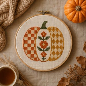 May include: A circular wooden embroidery hoop containing a cross-stitch design of a pumpkin on white fabric. The pumpkin features three vertical sections: a checkered orange pattern, a central floral motif, and a yellow diamond pattern.
