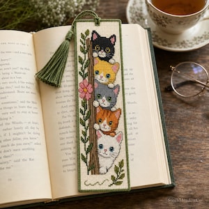 May include: A rectangular cross-stitch bookmark featuring a vertical stack of five kittens in black, yellow, gray, orange, and white colors climbing a brown tree trunk with green leaves and a pink flower, finished with a sage green fabric tassel.