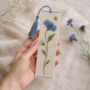 May include: A rectangular bookmark made of cream-colored Aida fabric featuring a cross-stitch design of a blue cornflower and a small bee. The bookmark is finished with a light blue braided cord and a matching tassel at the top edge.
