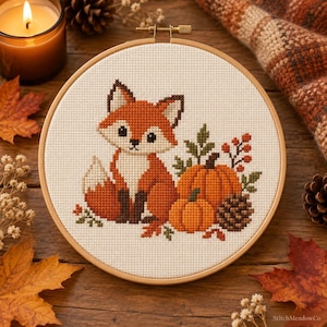 Op de afbeelding: Een ronde houten borduurring met een kruissteekpatroon van een kleine oranje en witte vos die naast twee oranje pompoenen, een bruine dennenappel en kleine groene blaadjes op een crèmekleurige stoffen achtergrond zit.