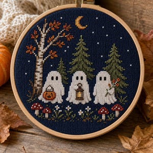 May include: A circular wooden embroidery hoop containing a cross-stitch design on dark blue fabric. The pattern features three white ghosts standing in a forest with pine trees, a birch tree, red mushrooms, a yellow crescent moon, and stars.