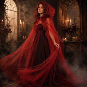 May include: A floor-length dark red strapless gown paired with a sheer, flowing red tulle hooded cape. The cape features a tie closure at the neck and drapes loosely over the shoulders, creating a voluminous silhouette with layers of fabric.