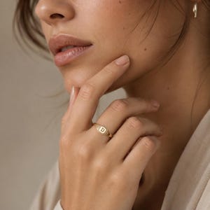 May include: A gold signet ring featuring a square face with a polished finish and the letter D engraved in the centre. The band is thin and smooth, crafted from a metallic gold material that reflects light across its surface.